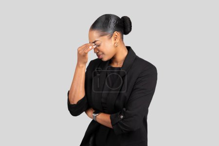 Téléchargez les photos : Une jeune femme afro-américaine dans un blazer noir touche son temple dans un geste de contemplation, les yeux fermés et l'expression sereine, isolée sur fond de studio gris - en image libre de droit