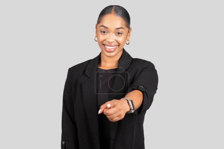 Foto de Involucrar a la joven afroamericana sonriente en una chaqueta negra a medida extiende su brazo con confianza hacia la cámara, sonriendo, motivada, aislada en un fondo gris del estudio - Imagen libre de derechos