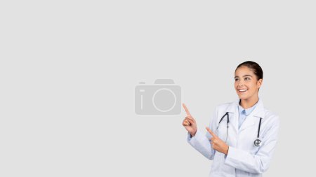 Téléchargez les photos : Souriant caucasien millénium dame médecin en manteau blanc, pointez du doigt l'espace de copie, recommander des soins de santé, isolé sur fond de studio gris. Médecine, publicité et offre, vente - en image libre de droit