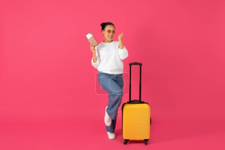 Téléchargez les photos : Joyeuse jeune voyageuse asiatique tenant un passeport avec des billets et célébrant le succès, heureuse femme debout près de la valise jaune sur fond rose, excitée pour le voyage à venir, espace de copie - en image libre de droit