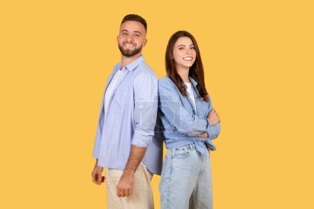 Téléchargez les photos : Souriant jeune homme et femme caucasien en tenue décontractée se tenir dos à dos, exsudant confiance et bonheur sur fond jaune vibrant - en image libre de droit