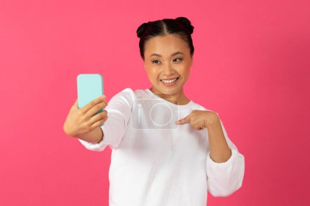 Foto de Feliz joven asiática mujer tomando selfie en el teléfono inteligente y apuntando a sí misma con expresión lúdica, alegre hermosa dama coreana posando en la cámara móvil, de pie sobre el fondo rosa - Imagen libre de derechos