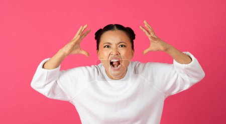 Foto de Furiosa joven mujer asiática en sudadera blanca gritando a la cámara, frustrada dama enojada levantando las manos con rabia, expresando emociones negativas, de pie aislada contra el vibrante fondo rosa del estudio - Imagen libre de derechos