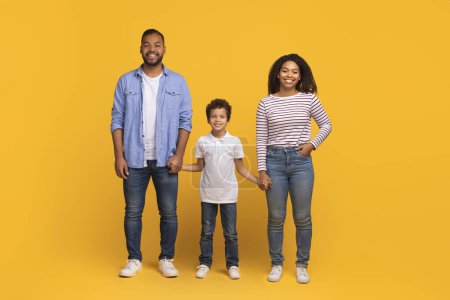 Téléchargez les photos : Famille noire confiante avec un petit fils debout main dans la main sur fond jaune, aimant les parents afro-américains et l'enfant mâle tenant la main et souriant à la caméra, dépeignant l'unité et le bonheur - en image libre de droit