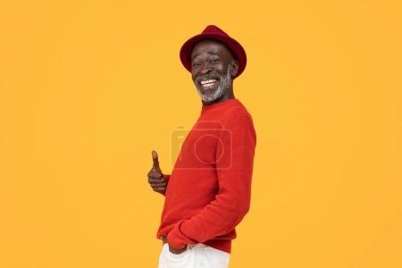 Photo for Cheerful confident senior african american man in a stylish red sweater and hat giving a thumbs-up sign, his broad smile expressing positivity and satisfaction on a yellow background, studio - Royalty Free Image