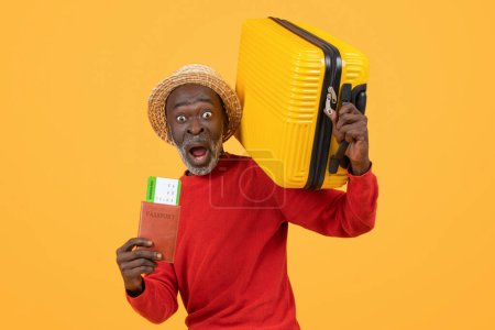 Foto de Hombre afroamericano mayor sobresaltado con un sombrero de paja y suéter rojo con pasaporte con tarjeta de embarque y una maleta amarilla, con una expresión sorprendida sobre un fondo amarillo - Imagen libre de derechos