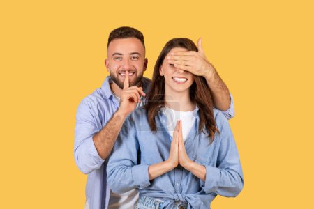 Photo for Playful man is placing his finger to his lips for silence while covering the eyes of smiling woman, both in blue shirts on yellow background - Royalty Free Image