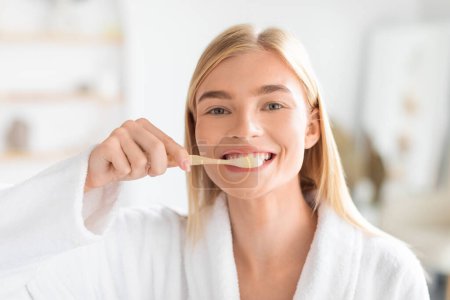 Foto de Mujer rubia joven cepillándose los dientes centrándose en la higiene bucal como parte de la rutina matutina en un baño moderno. Retrato de mujer con cepillo de dientes cuidando sus dientes limpiando - Imagen libre de derechos