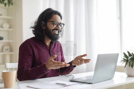 Online Tutoring. Young indian tutor wearing glasses and headset making video call on laptop, smiling male teacher hand gesturing while explaining topic in online lesson, working from his home office
