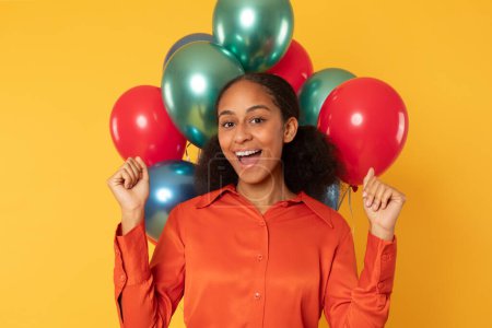 Photo for Holiday party. Happy African American teenager girl raising hands in victorious gesture, posing among festive balloons over yellow backdrop. Studio shot of joyful teen lady celebrating birthday - Royalty Free Image