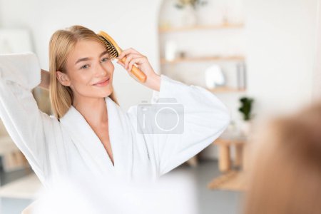 Photo for Blonde young lady brushes hair smiling at mirror reflection in modern bathroom, representing beauty routine and selfcare. Woman using hairbrush styling her hair after shower - Royalty Free Image