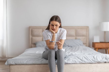 Photo for Sad disappointment lady holding pregnancy test, sitting on bed alone in bedroom interior. Infertility, female health problems, unwanted pregnancy and loneliness after breakup - Royalty Free Image