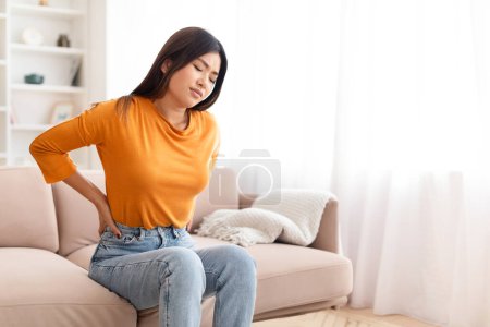 Photo for Unhappy young chinese woman have back pain after nap on couch at home, massaging lower back with both hands, copy space. Bad posture problem, osteochondrosis - Royalty Free Image