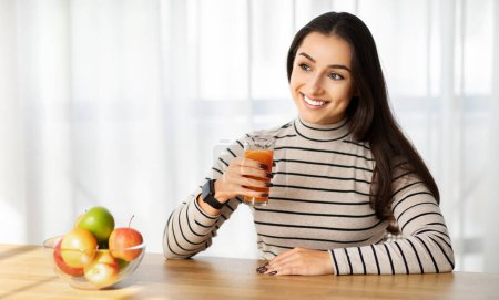 Téléchargez les photos : Joyeux jeune femme du Moyen-Orient profitant d'un verre de jus frais, avec un bol de fruits colorés sur la table, la promotion d'un mode de vie sain et la nutrition à la maison cuisine - en image libre de droit