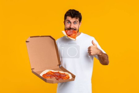Photo for Happy Guy With Slice Of Pizza In Mouth Gesturing Thumbs Up On Yellow Studio Background, Approving Pizzeria Delivery And Taste, Smiling To Camera. Great Junk Food Concept, Cheat Meal - Royalty Free Image