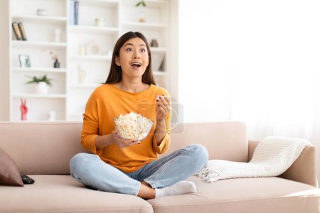 Téléchargez les photos : Incroyable jeune femme asiatique regardant un film intéressant ou un programme de télévision à la maison, assis sur le canapé dans un salon confortable, mangeant du pop-corn, profitant d'un bon film. Divertissement domestique, espace de copie - en image libre de droit