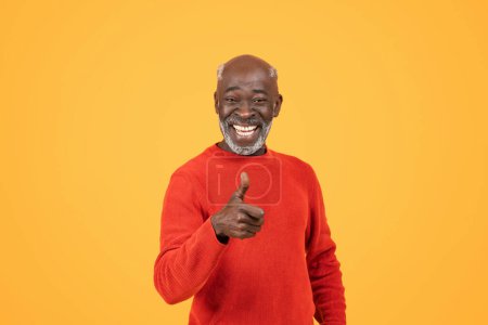 Photo for Smiling senior man with a vibrant personality gives a thumbs-up sign while wearing a red sweater and hat against an energetic yellow background - Royalty Free Image