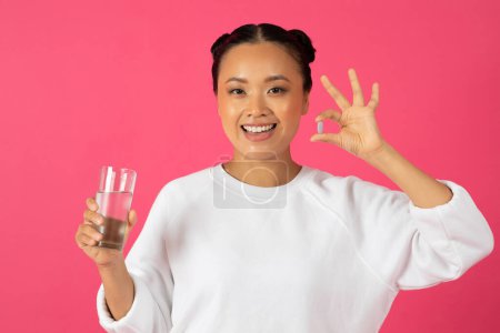 Foto de Nutrición Dieta Saludable. Mujer asiática sonriente sosteniendo la píldora de vitamina y un vaso de agua, Feliz Joven coreano femenino recomendando suplementos de belleza, de pie aislado sobre fondo rosa, espacio de copia - Imagen libre de derechos