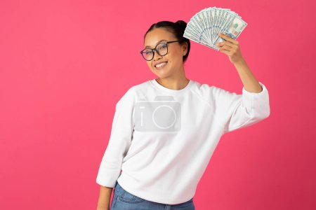 Foto de Gran beneficio. Mujer asiática alegre con gafas sosteniendo ventilador de billetes de dólar, señora feliz mostrando dinero en efectivo, significando éxito financiero, de pie aislado en el fondo del estudio rosa, espacio de copia - Imagen libre de derechos