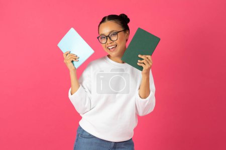 Photo for Cheerful asian woman wearing glasses holding books in hands and smiling at camera, happy young korean student lady enjoying education, standing isolated against bright pink background, copy space - Royalty Free Image