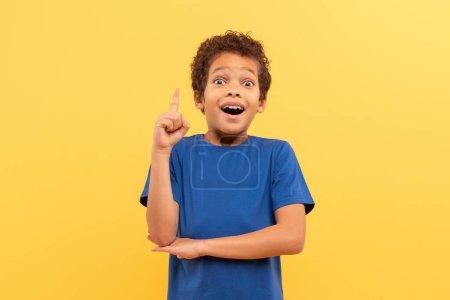 Téléchargez les photos : Surpris jeune garçon aux cheveux bouclés, ayant un moment aha, pointant vers le haut avec une expression large, en chemise bleue sur fond jaune, incarnant une inspiration soudaine - en image libre de droit