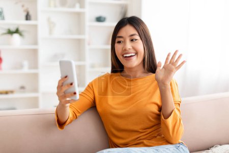 Foto de Positiva mujer asiática bastante joven sentada en el sofá en casa, saludando y sonriendo en la pantalla del teléfono inteligente, tienen videollamada con familiares o amigos, espacio para copiar, concepto de telecomunicaciones - Imagen libre de derechos
