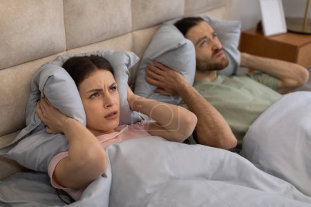 Téléchargez les photos : Jeune couple caucasien souffrant de voisins bruyants, couvrant les oreilles avec des oreillers tout en dormant au lit la nuit à la maison. Concept d'appartement bruyant - en image libre de droit