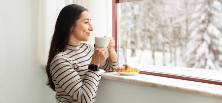 Photo for Serene smiling middle eastern millennial woman in striped turtleneck savoring the taste of her morning coffee by a snowy window, her expression peaceful and a croissant on the sill - Royalty Free Image