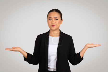 Photo for Perplexed Asian businesswoman with palms up in a questioning gesture, showing uncertainty or confusion, in a professional black blazer and white shirt, against a light grey background - Royalty Free Image