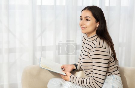 Téléchargez les photos : Une jeune femme souriante du Moyen-Orient au col roulé rayé s'assoit confortablement sur un canapé, tenant un stylo et un carnet, prenant peut-être des notes ou un journal dans une pièce lumineuse et aérée - en image libre de droit