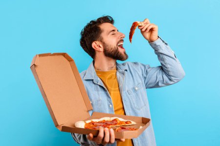 Photo for Pizza Lover. Funny Man Overeating Junk Food, Eating Slice Of Pizza Posing With Box Against Blue Studio Background, Showcasing Unhealthy Nutrition Habits. Guy Enjoys Cheat Meal Concept - Royalty Free Image