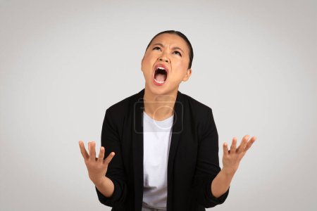 Photo for Desperate Asian businesswoman with an anguished expression, hands up in the air, looking overwhelmed and stressed, wearing a black blazer and white shirt on a grey background - Royalty Free Image