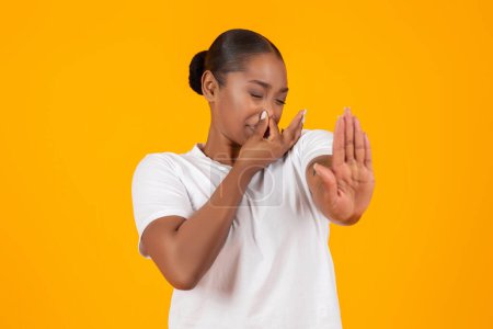 Téléchargez les photos : Mauvaise odeur. Jeune femme noire dégoûtée fermant le nez et montrant STOP geste, exprimant dégoût sur fond jaune studio. Femme millénaire repoussée par une odeur toxique ou un parfum terrible - en image libre de droit