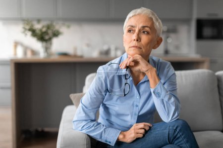 Photo for Depression. Portrait of unhappy european mature lady with gray short hair, looking aside with upset expression, holding eyeglasses while thinking about her problems seated on sofa at home - Royalty Free Image