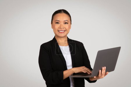 Foto de Una joven empresaria asiática confiada en un blazer negro y una blusa blanca sostiene un portátil abierto en sus manos, sonriendo a la cámara sobre un fondo gris claro. Negocios y trabajo - Imagen libre de derechos