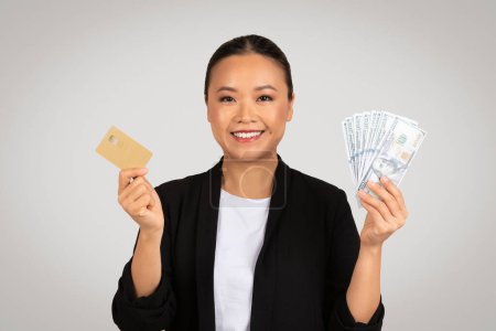 Téléchargez les photos : Souriante femme d'affaires asiatique affiche avec confiance à la fois la carte de crédit dorée et fan de dollars américains, symbolisant la richesse, les choix financiers et le pouvoir d'achat, isolé sur fond gris, studio - en image libre de droit
