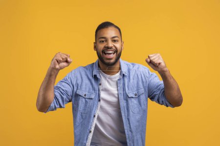 Photo for Joyful young black man celebrating success on yellow studio background, happy excited african american male shaking fists, handsome guy cheering luck and sharing good emotions, copy space - Royalty Free Image