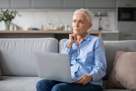 Photo for Worried senior lady using laptop and touching chin thinking about issues, sitting with computer on sofa modern living room interior. Retirement financial problems and stress - Royalty Free Image