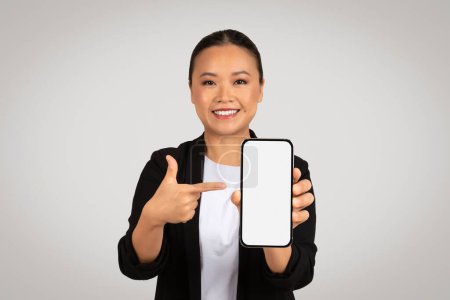 Photo for Confident Asian businesswoman presenting a smartphone with a blank white screen, pointing with her index finger, expressing positivity and approval, isolated on gray background, studio - Royalty Free Image