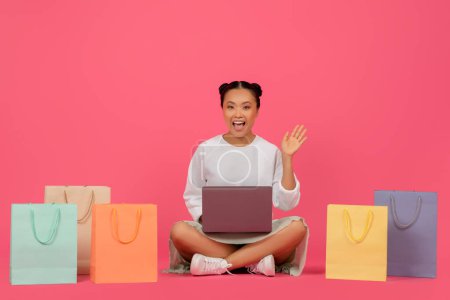 Foto de Gran sitio web. Mujer asiática emocionada con el ordenador portátil sentado entre las bolsas de compras, Happy Korean Lady Shopping Online, Disfrutando de hacer compras por Internet, Posando en el fondo del estudio rosa, Copiar el espacio - Imagen libre de derechos