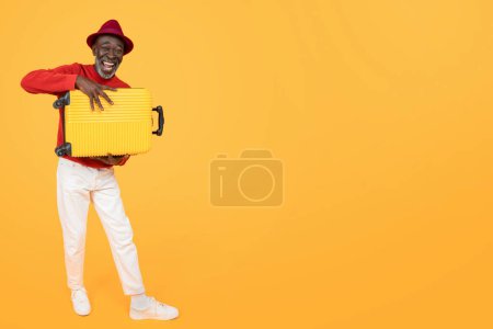 Foto de Hombre afroamericano alegre mayor en un suéter rojo y sombrero rojo de moda que lleva una maleta de color amarillo brillante, con una sonrisa radiante, listo para viajar en un estudio de fondo amarillo - Imagen libre de derechos