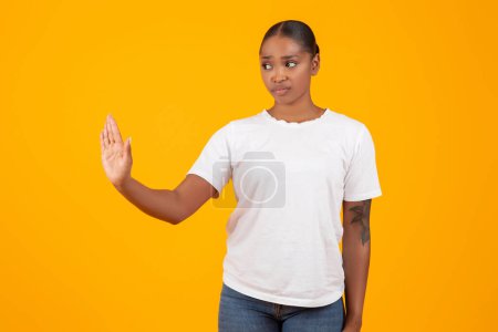 Téléchargez les photos : Offensée femme noire millénaire en t-shirt blanc fait geste d'arrêt avec la main sur fond de studio jaune, rejetant la mauvaise offre et exprimant sa désapprobation. Lutte contre le harcèlement, le sexisme - en image libre de droit