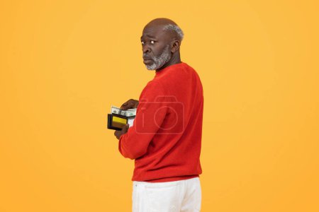 Photo for Skeptical sad elderly African American man in a red sweater discreetly holding a wallet full of cash over his shoulder on a yellow background, looking wary or cautious - Royalty Free Image