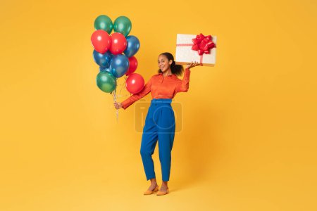 Téléchargez les photos : Pleine longueur de jeune fille noire heureuse célébrant anniversaire tenant tas de ballons colorés et boîte cadeau avec cadeau, debout sur fond jaune, espace libre, endroit pour la publicité - en image libre de droit