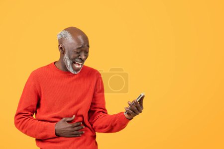 Foto de Alegre anciano alegre en un suéter rojo se ríe mientras mira un teléfono inteligente, mano en el estómago, de pie sobre un fondo amarillo vibrante, lo que significa buenas noticias o una broma - Imagen libre de derechos