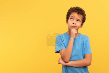 Téléchargez les photos : Jeune garçon réfléchi aux cheveux bouclés regardant de côté l'espace de copie, touchant le menton en contemplation, portant une chemise bleue sur fond jaune, incarnant la curiosité et la pensée profonde - en image libre de droit