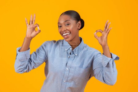 Téléchargez les photos : Jeune femme noire souriante montrant le geste OKAY avec les deux mains sur fond de studio jaune. Dame afro-américaine joyeuse exprimant son approbation et donnant un retour positif - en image libre de droit