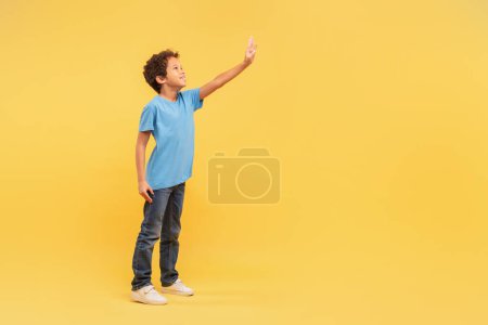 Photo for Inquisitive young boy with curly hair reaching out with his hand at free space, guy wearing blue t-shirt on yellow background, symbolizing curiosity and exploration - Royalty Free Image