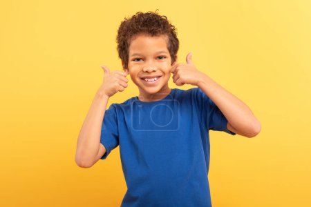 Foto de Niño alegre con el pelo rizado dando pulgares cercanos hacia arriba por su cara, con amplia sonrisa, en camiseta azul real, sobre fondo amarillo mostaza, señalando aprobación y felicidad - Imagen libre de derechos