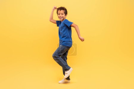 Foto de Muchacho exuberante con el pelo rizado bailando alegremente en camiseta azul y jeans sobre fondo amarillo brillante, expresando felicidad y energía - Imagen libre de derechos
